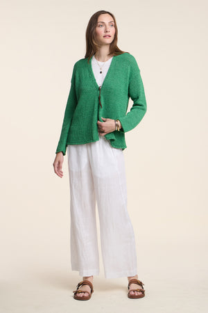 A woman stands against a plain background wearing a green sweater, brown sandals, layered jewelry, and the LOLA LINEN PANT by Velvet by Graham & Spencer, with her hands relaxed at her sides.