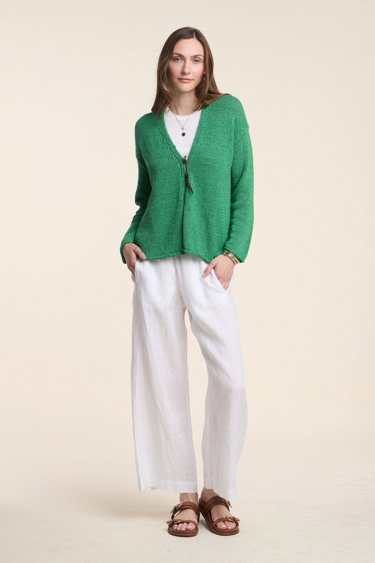 A woman faces forward in the QUINNLY CARDIGAN by Velvet by Graham & Spencer, a green open-front style with long drop shoulders, layered over a white top and wide-legged white pants, finished with brown sandals and layered necklaces.-41527107387585