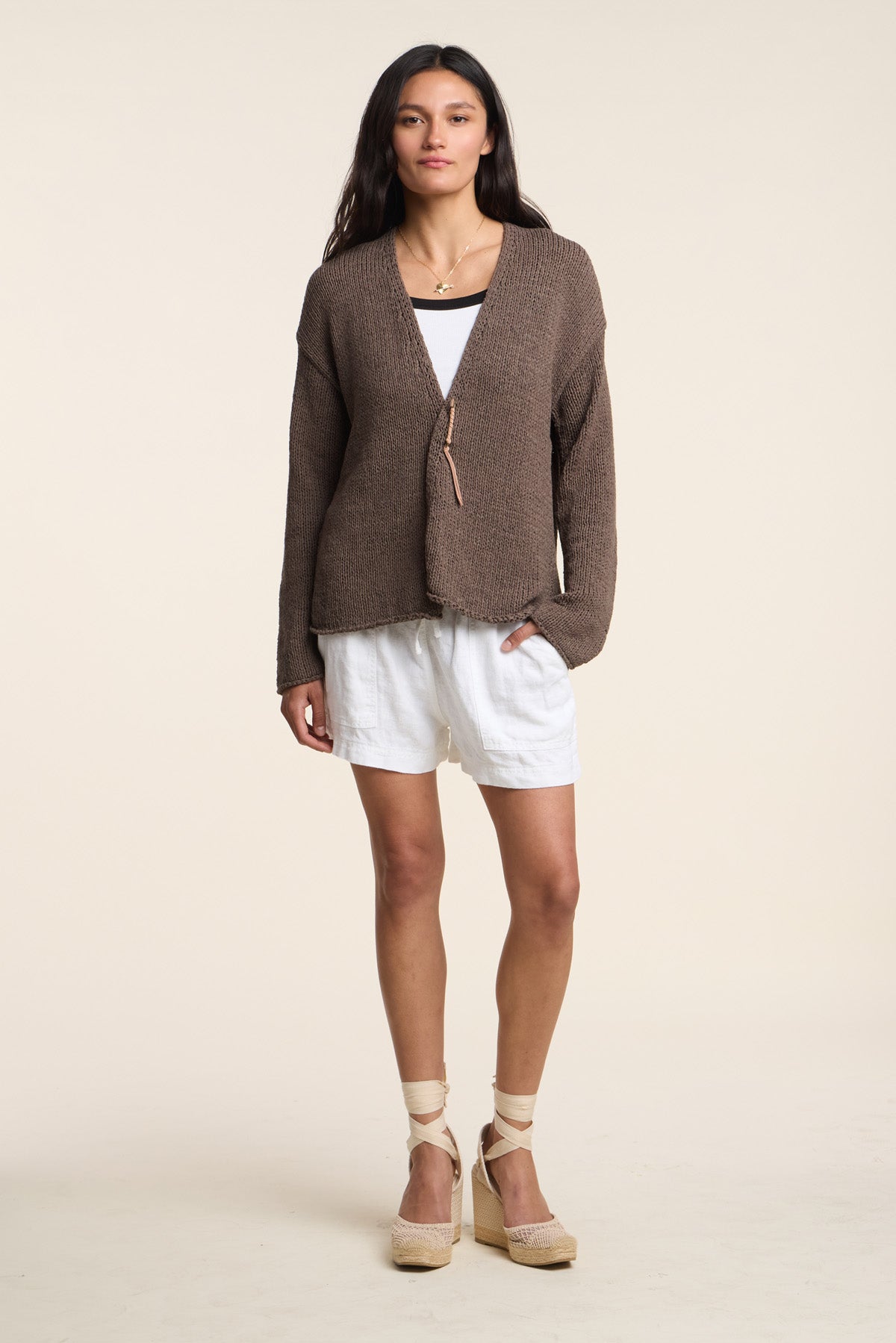 A woman faces forward against a plain background wearing the QUINNLY CARDIGAN by Velvet by Graham & Spencer, styled with a white tank top, white shorts, and beige wedge sandals.-41527107518657