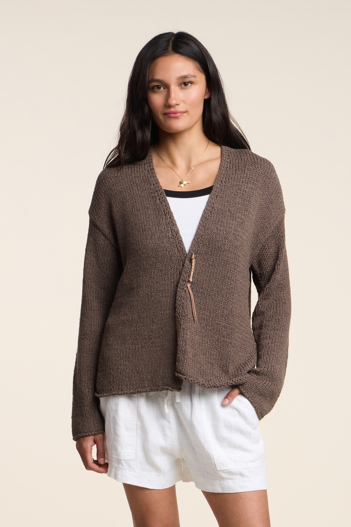A person wears the QUINNLY CARDIGAN by Velvet by Graham & Spencer—an open-front cotton tape yarn cardigan—over a white top and shorts, standing with one hand in their pocket against a plain background.