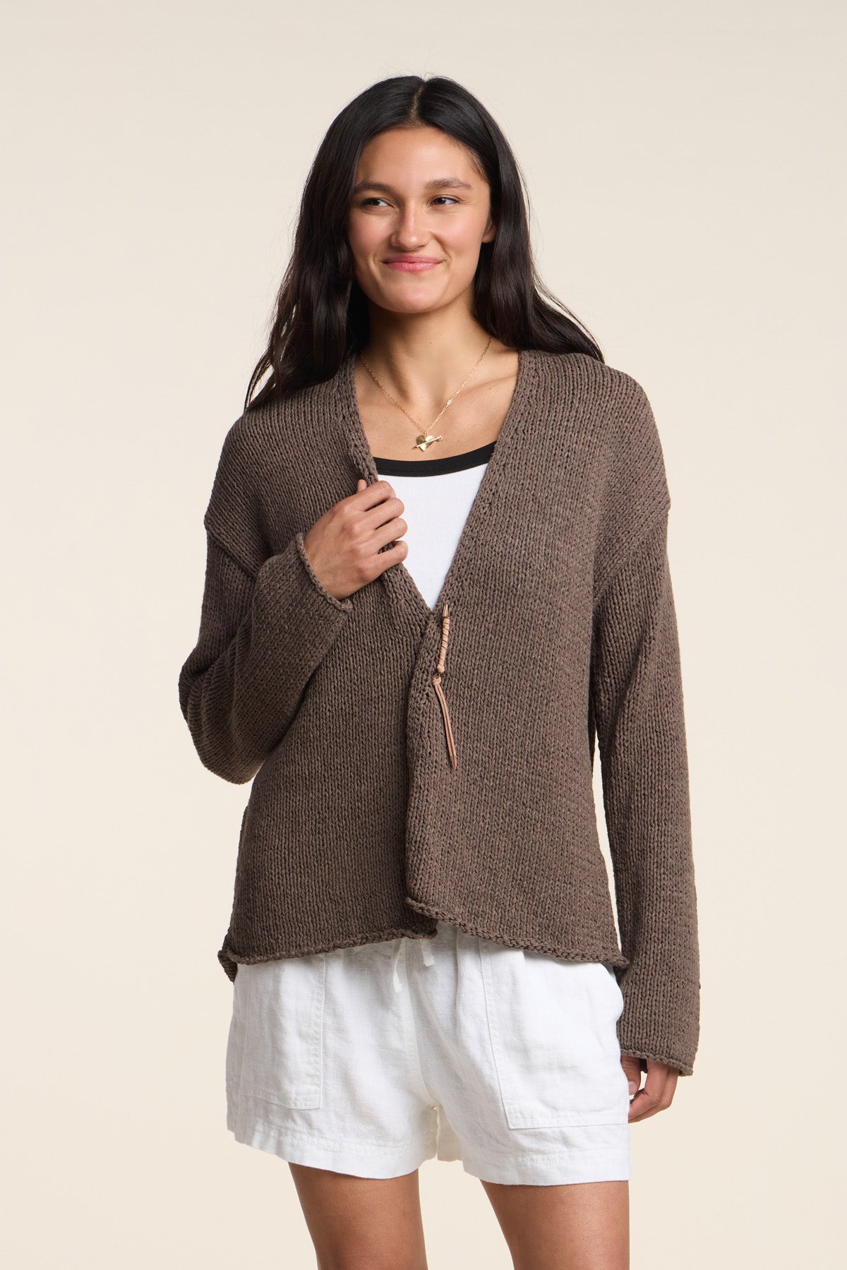 A person wears the QUINNLY CARDIGAN by Velvet by Graham & Spencer—an open-front brown cotton tape yarn piece—over a white top and shorts, standing against a plain background and smiling slightly.