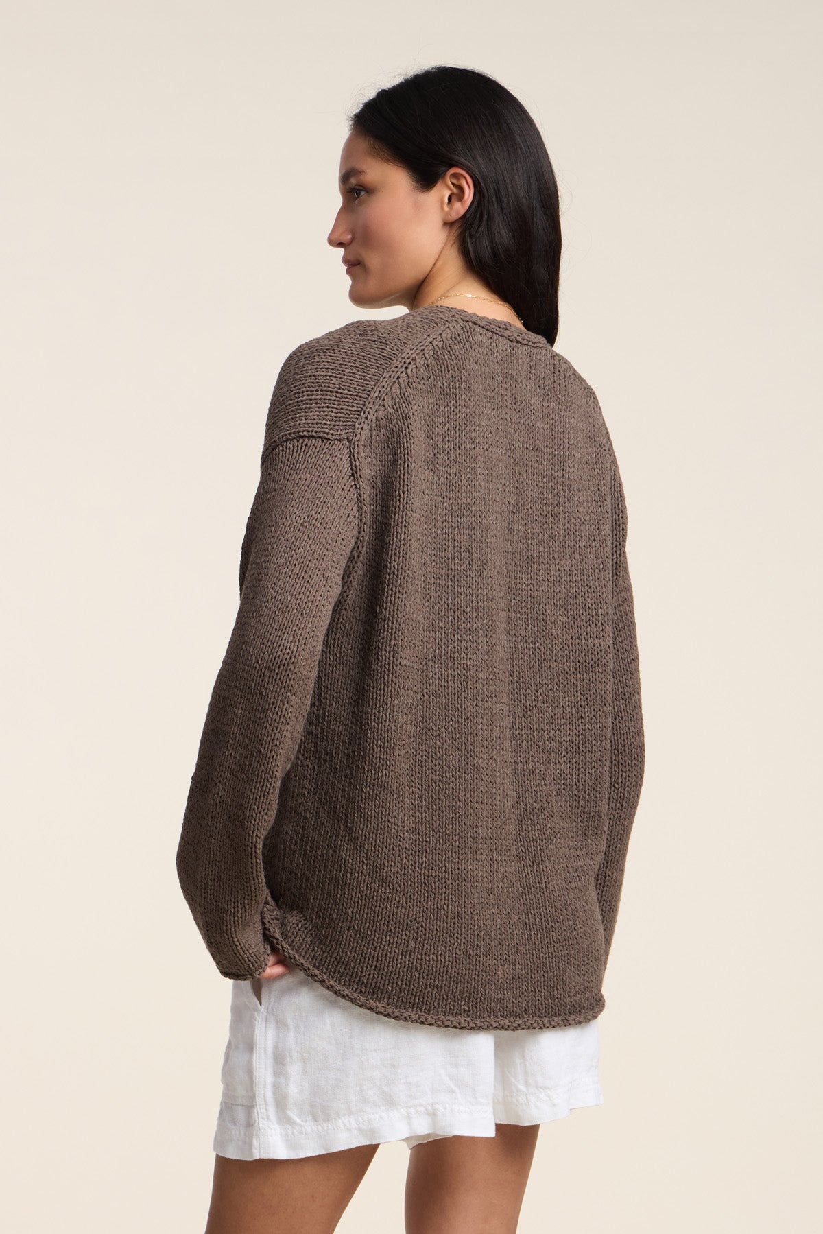 A woman stands with her back to the camera, wearing the QUINNLY CARDIGAN by Velvet by Graham & Spencer, a brown open-front cotton tape yarn cardigan, paired with white shorts against a neutral background.