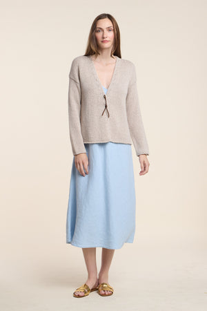 A woman stands against a plain background wearing a light blue midi dress, gold sandals, and the QUINNLY CARDIGAN by Velvet by Graham & Spencer—a beige open-front style with a tie front, crafted from cotton tape yarn.