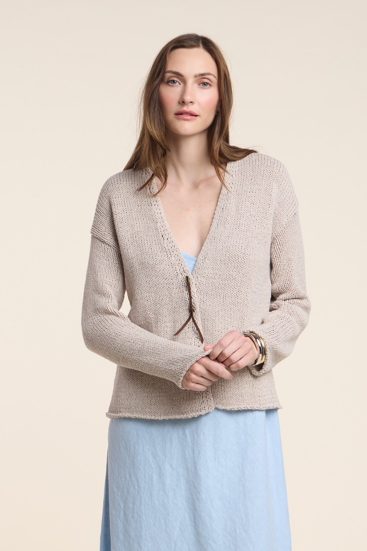 A woman wearing the QUINNLY CARDIGAN by Velvet by Graham & Spencer, a beige open-front piece knit from soft cotton tape yarn, stands against a light background over a light blue dress, looking at the camera with her hands clasped.