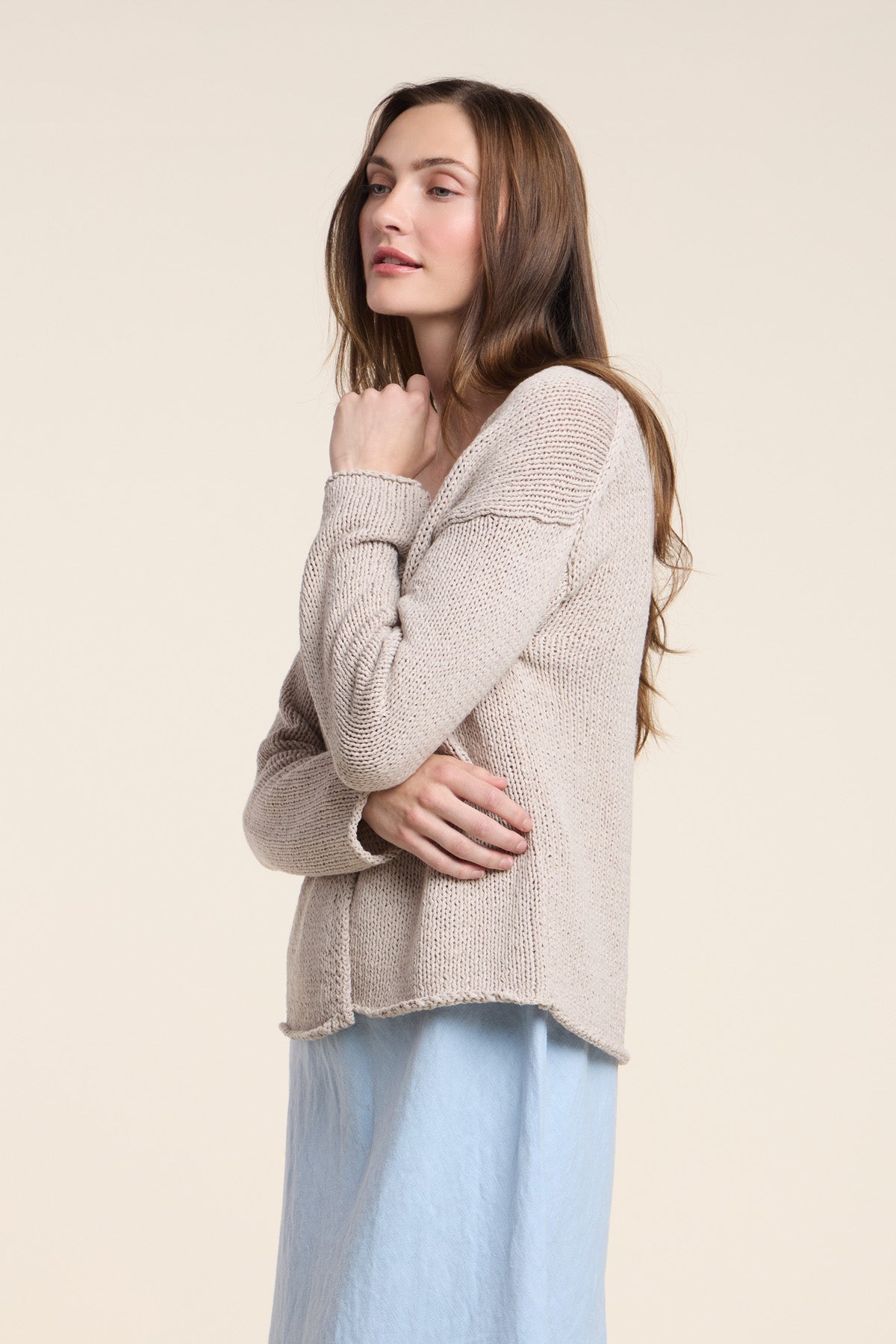 A woman with long brown hair stands in profile against a plain light background, wearing the QUINNLY CARDIGAN by Velvet by Graham & Spencer—an open-front beige cardigan crafted from cotton tape yarn—paired with a light blue skirt.