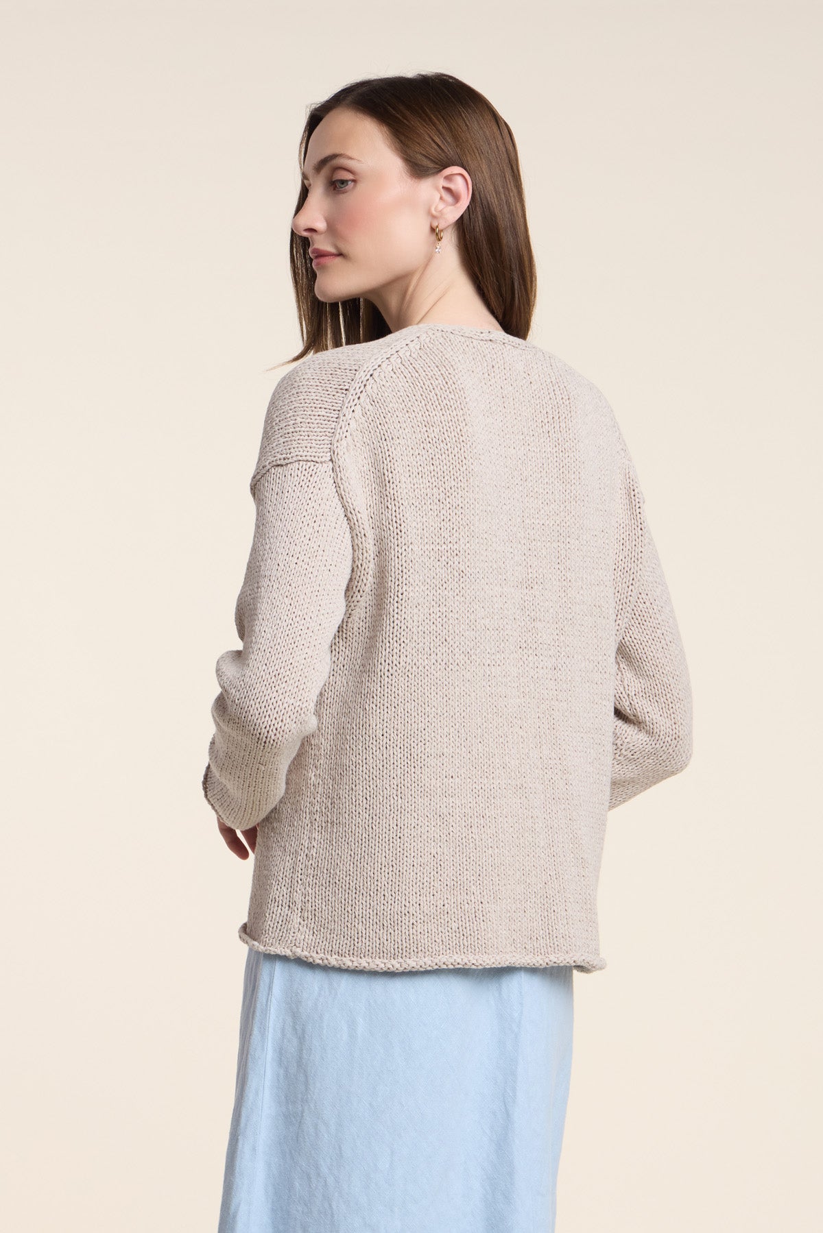 A woman in the QUINNLY CARDIGAN by Velvet by Graham & Spencer, a beige knit sweater, pairs it with a light blue skirt and stands facing away, glancing over her shoulder against a neutral background.