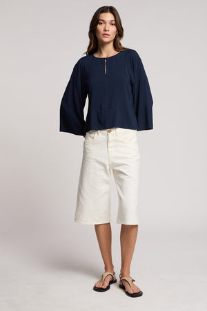 A woman faces forward wearing the navy RAEGAN TOP by Velvet by Graham & Spencer, paired with knee-length white shorts and black sandals against a plain background.