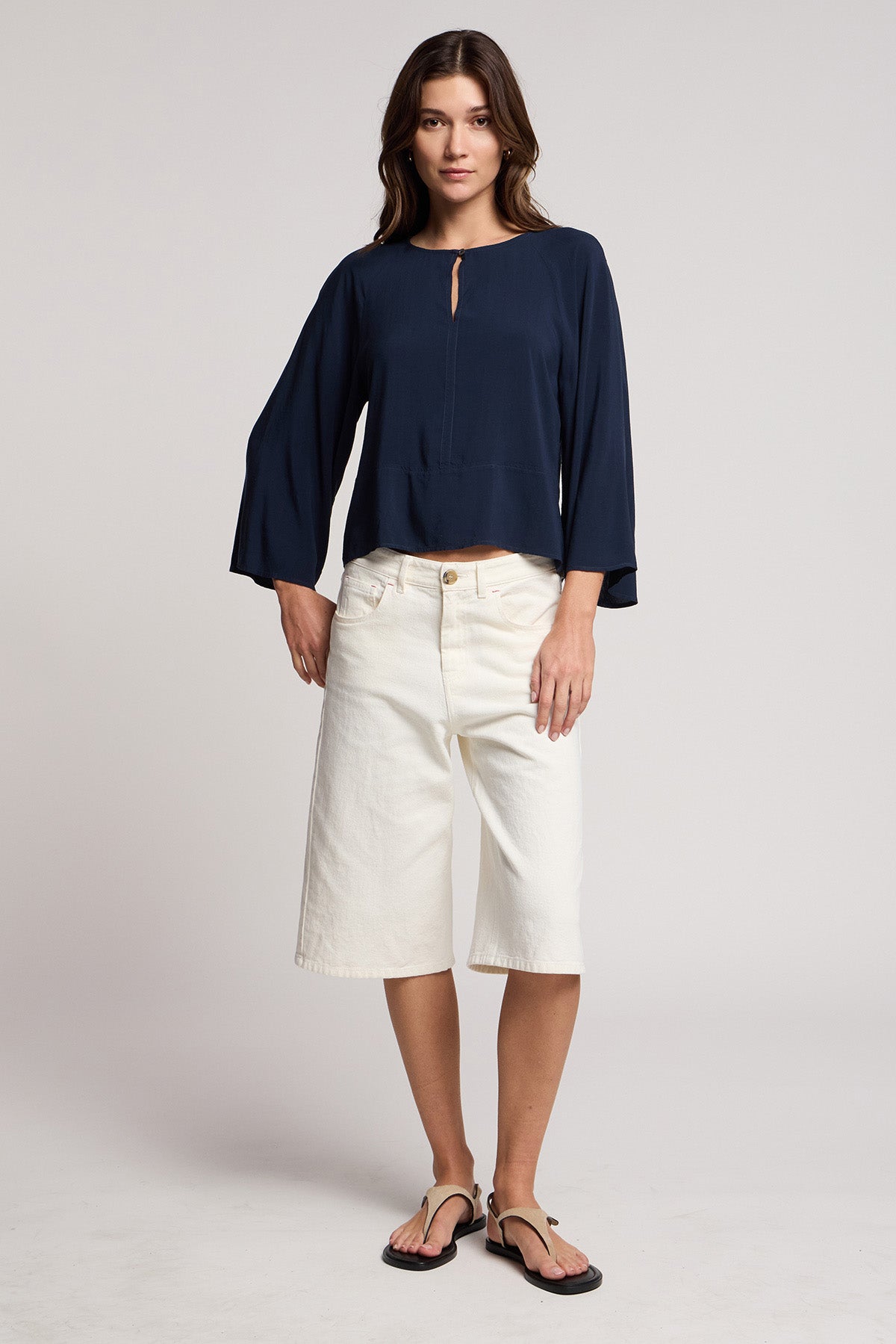 A woman stands facing forward wearing the OAKLEY DENIM SHORT by Velvet by Graham & Spencer, paired with a navy long-sleeve blouse and black flip-flops against a plain background.-41371966210241