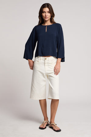 A woman stands facing forward wearing the OAKLEY DENIM SHORT by Velvet by Graham & Spencer, paired with a navy long-sleeve blouse and black flip-flops against a plain background.