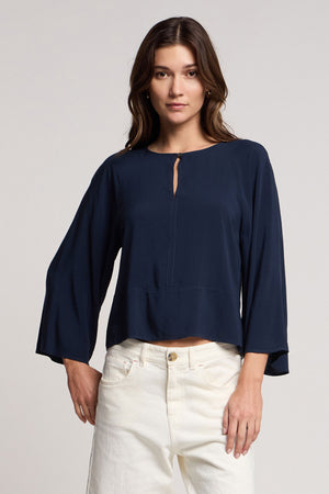 A woman wears the Velvet by Graham & Spencer RAEGAN TOP—a long-sleeve, navy rayon challis blouse with bell sleeves and a front keyhole—paired with white pants against a plain background.