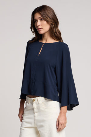 A woman stands against a plain background, wearing the Velvet by Graham & Spencer RAEGAN TOP in soft rayon challis with wide bell sleeves, paired with off-white pants.