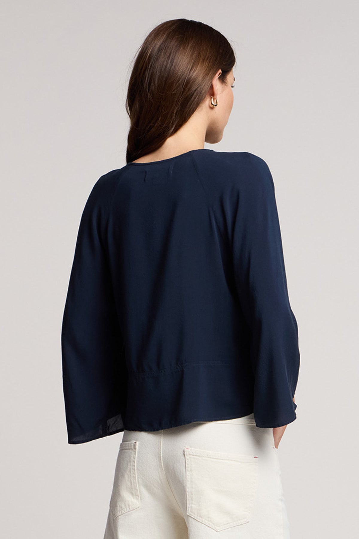 A woman with long brown hair is seen from the back against a plain background, wearing Velvet by Graham & Spencer’s RAEGAN TOP in navy with bell sleeves and white pants.-41371967488193