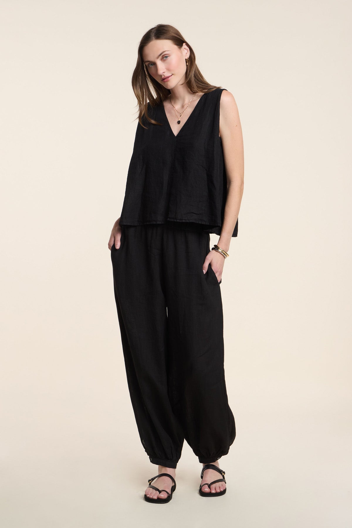 A woman wears the RENNA LINEN TOP by Velvet by Graham & Spencer—a loose black sleeveless summer top—paired with black baggy pants and sandals, standing against a plain background.-41371967750337
