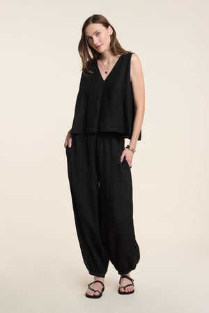 A woman wears the RENNA LINEN TOP by Velvet by Graham & Spencer—a loose black sleeveless summer top—paired with black baggy pants and sandals, standing against a plain background.