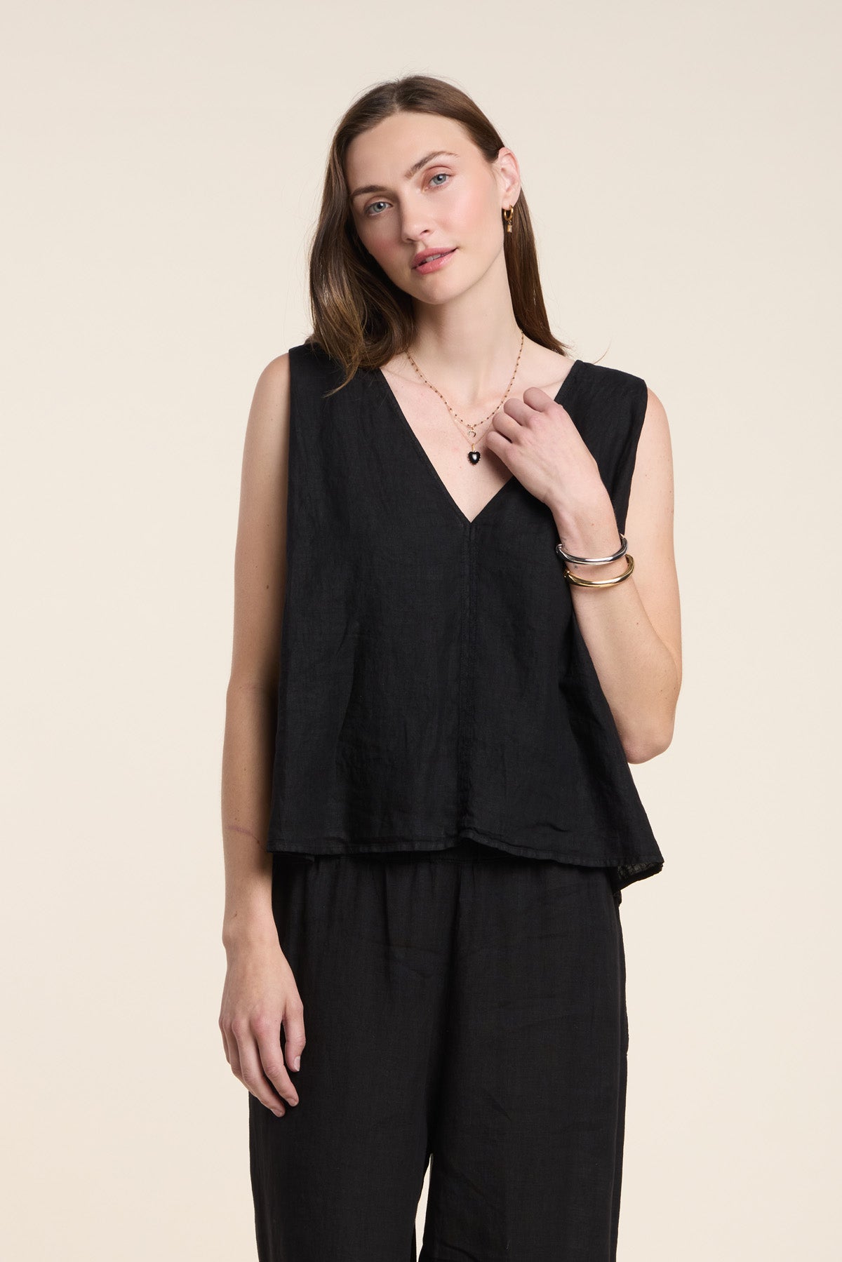 A woman stands against a plain background wearing the RENNA LINEN TOP by Velvet by Graham & Spencer, a sleeveless V-neck summer blouse paired with black pants, minimal jewelry, and a relaxed expression.