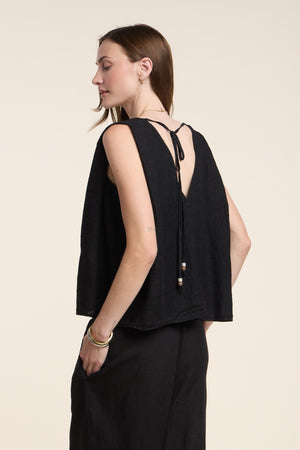 A woman stands sideways against a plain light background wearing the RENNA LINEN TOP by Velvet by Graham & Spencer—a sleeveless summer top featuring a V-shaped back with tie detail—paired with black pants.