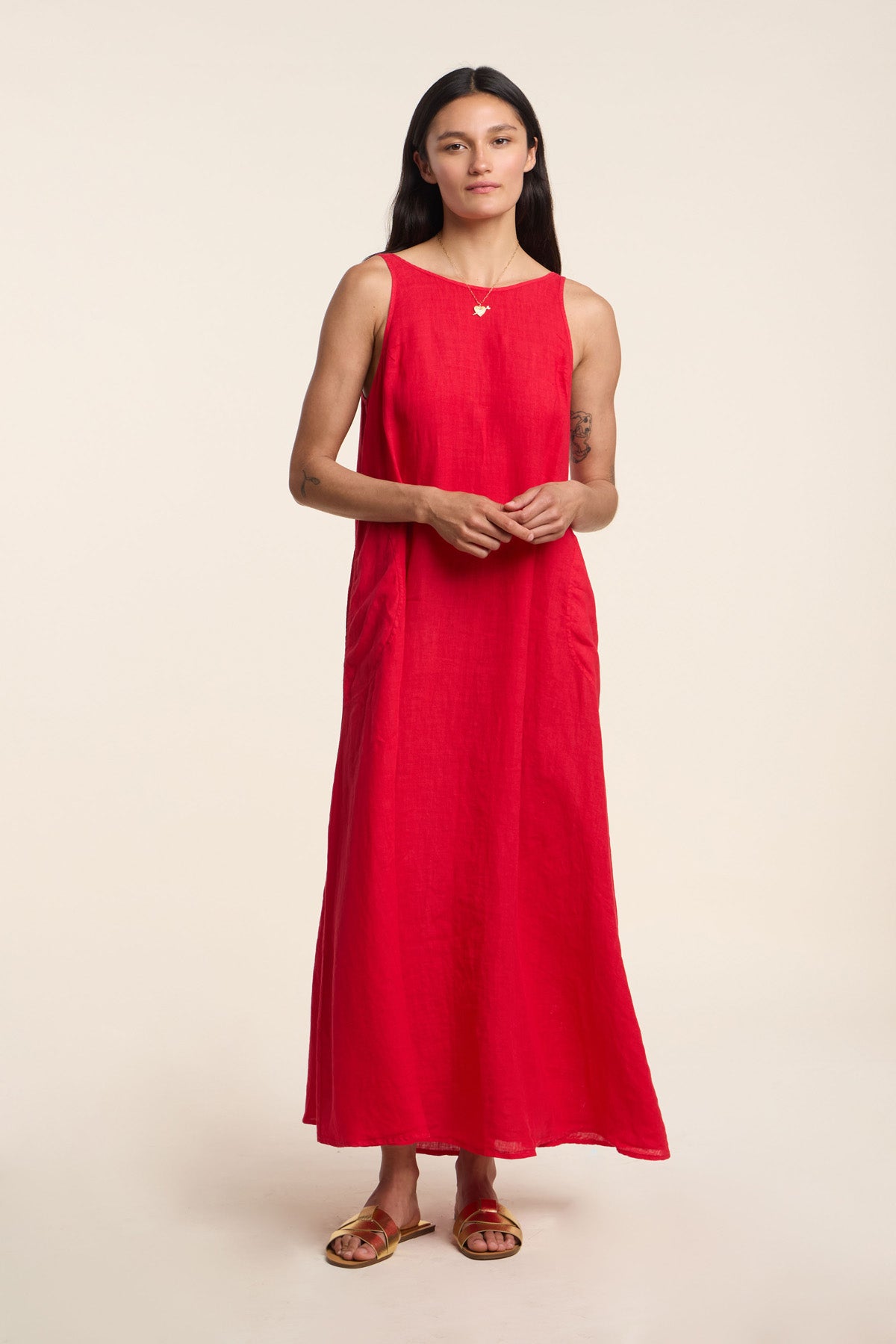 A woman wears the RETA LINEN DRESS by Velvet by Graham & Spencer—a sleeveless red maxi styled with sandals and a necklace, set against a plain light background for an effortlessly chic warm-weather look.-41372105867457