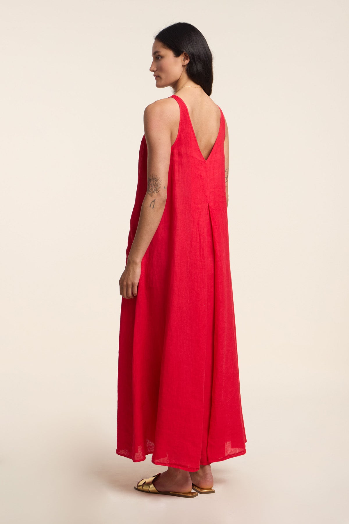 A woman stands sideways in the RETA LINEN DRESS by Velvet by Graham & Spencer—a loose, sleeveless red maxi dress with a deep V-back and gold sandals—showcasing effortless warm-weather style against a plain light background.-41372105965761