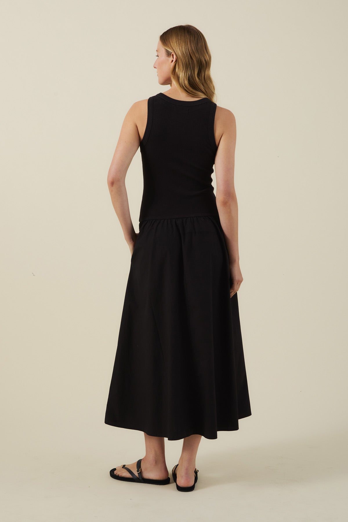 A woman stands facing away, wearing the ROMILLY DRESS by Velvet by Graham & Spencer— a black midi dress with a fitted ribbed bodice—paired with black sandals against a plain light background.-41527504044225