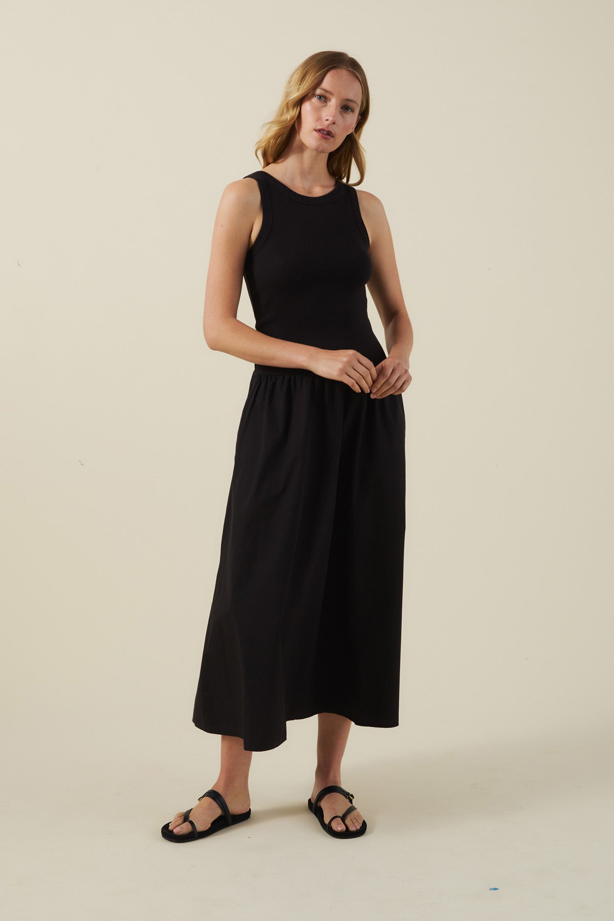 A woman stands against a plain background in the ROMILLY DRESS by Velvet by Graham & Spencer—a black cotton poplin dress with a fitted ribbed bodice, ankle-length skirt, and paired with black sandals.-41527504109761