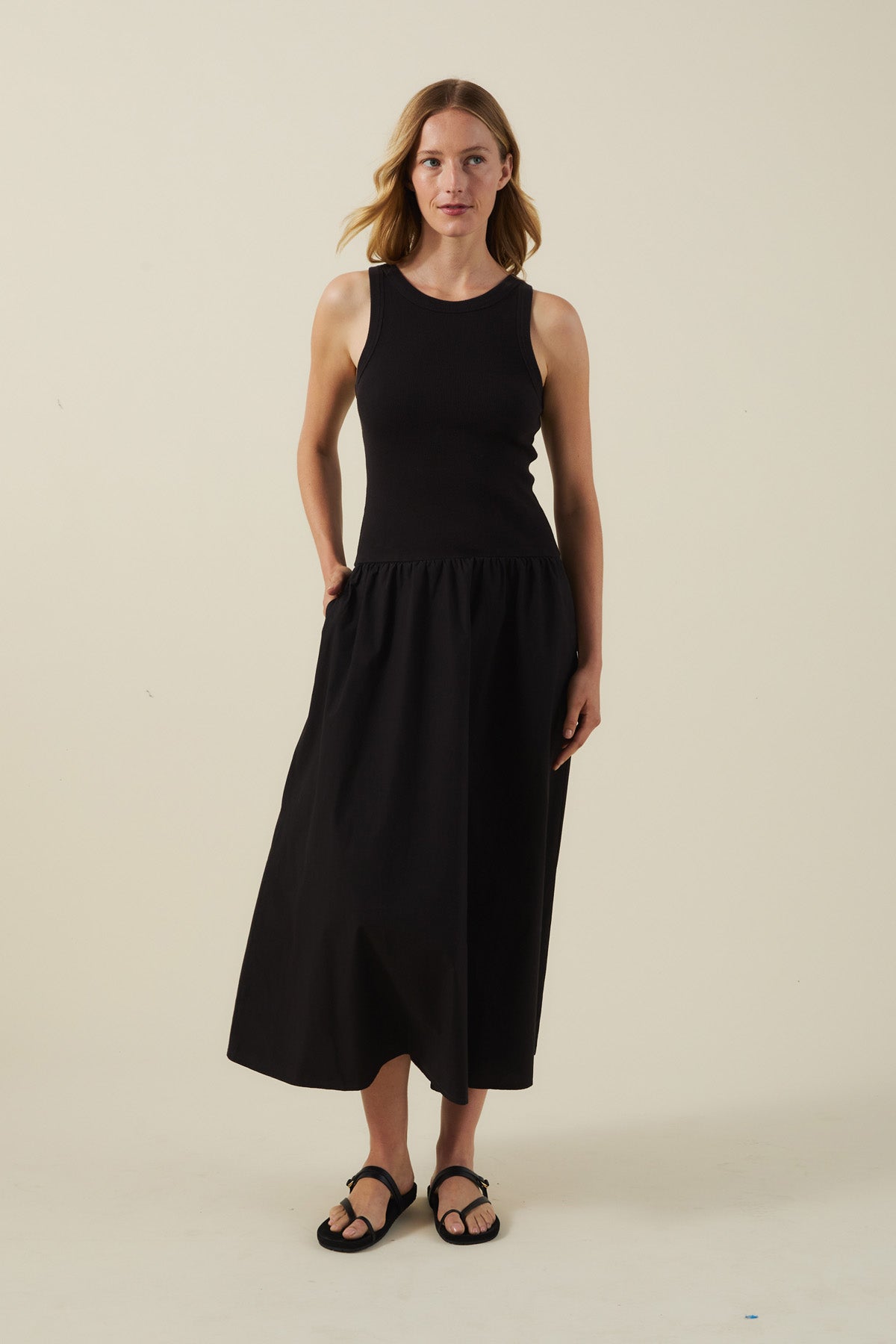 A woman stands against a plain background wearing the ROMILLY DRESS by Velvet by Graham & Spencer—a sleeveless black dress with a fitted ribbed bodice and cotton poplin skirt—paired with sandals.-41527504142529