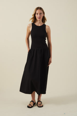 A woman stands against a plain background wearing the ROMILLY DRESS by Velvet by Graham & Spencer—a sleeveless black dress with a fitted ribbed bodice and cotton poplin skirt—paired with sandals.