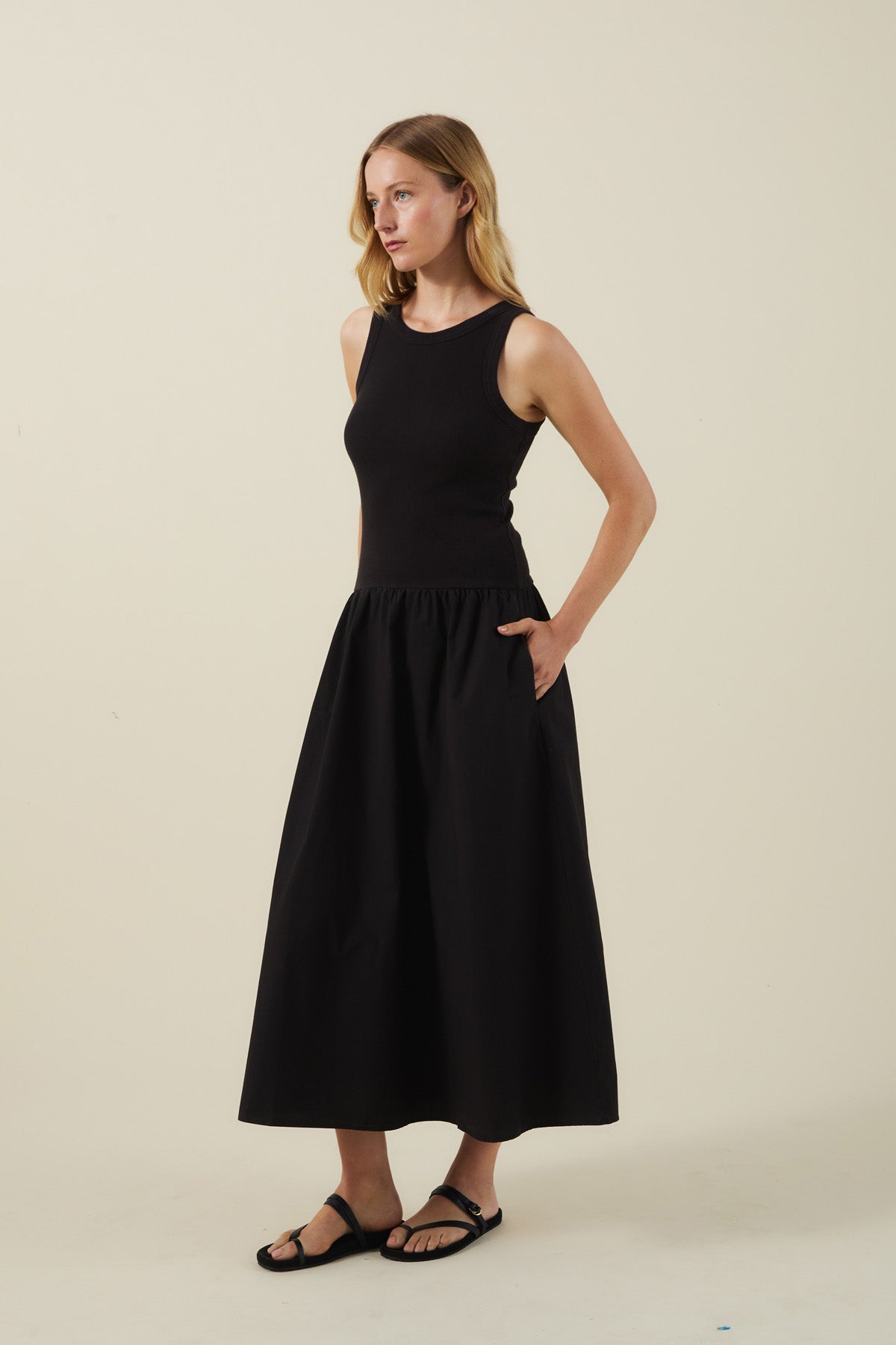 A woman stands against a plain background wearing the ROMILLY DRESS by Velvet by Graham & Spencer, a sleeveless black cotton poplin dress with pockets, styled with black sandals.