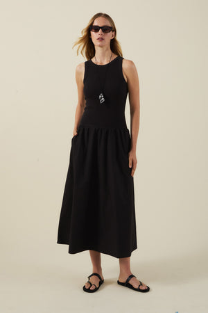 A woman wearing sunglasses, black sandals, a long necklace, and the ROMILLY DRESS by Velvet by Graham & Spencer stands against a plain light background.
