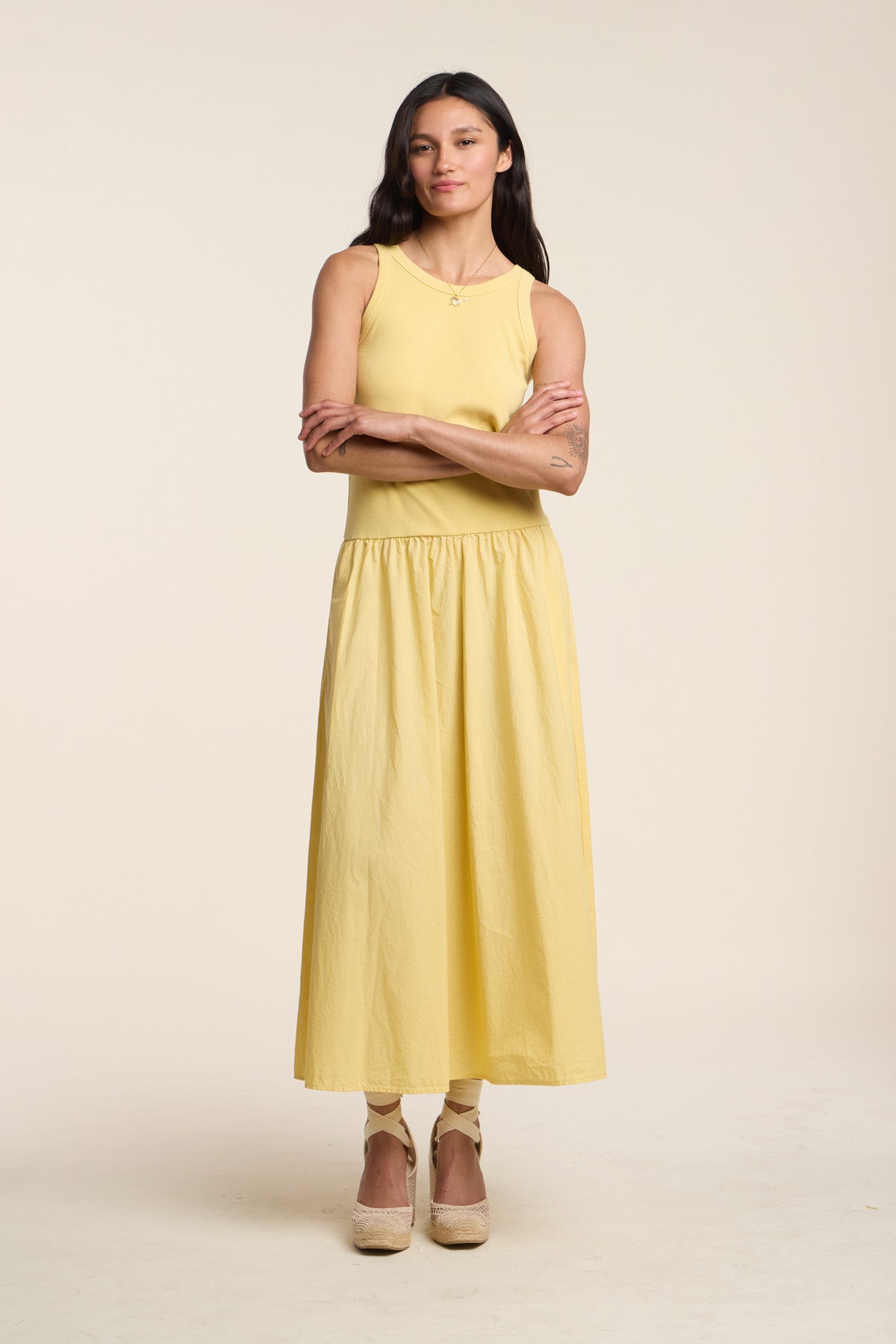 A woman stands with arms crossed, wearing the ROMILLY DRESS by Velvet by Graham & Spencer—a sleeveless yellow ribbed bodice dress—paired with beige platform shoes against a plain light background.