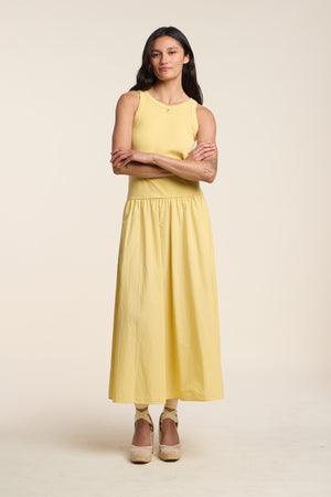 A woman stands with arms crossed, wearing the ROMILLY DRESS by Velvet by Graham & Spencer—a sleeveless yellow ribbed bodice dress—paired with beige platform shoes against a plain light background.