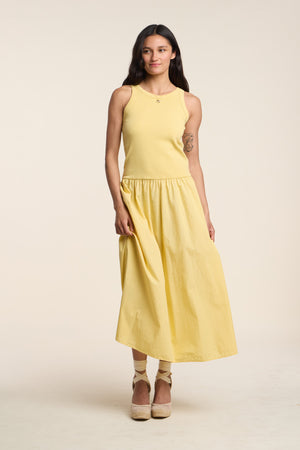 A woman stands against a plain background wearing the ROMILLY DRESS by Velvet by Graham & Spencer—a sleeveless yellow ribbed bodice dress with a matching cotton poplin skirt—and beige platform shoes.
