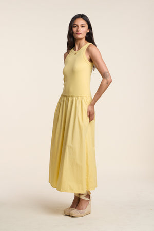 A woman stands against a plain background wearing the ROMILLY DRESS by Velvet by Graham & Spencer—a sleeveless yellow ribbed bodice dress with a matching cotton poplin skirt and beige wedge shoes.
