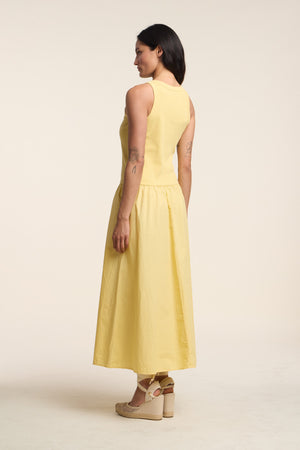 A woman faces away, wearing the ROMILLY DRESS by Velvet by Graham & Spencer—a yellow ribbed bodice dress with a flared cotton poplin skirt—paired with beige heeled sandals against a light background.