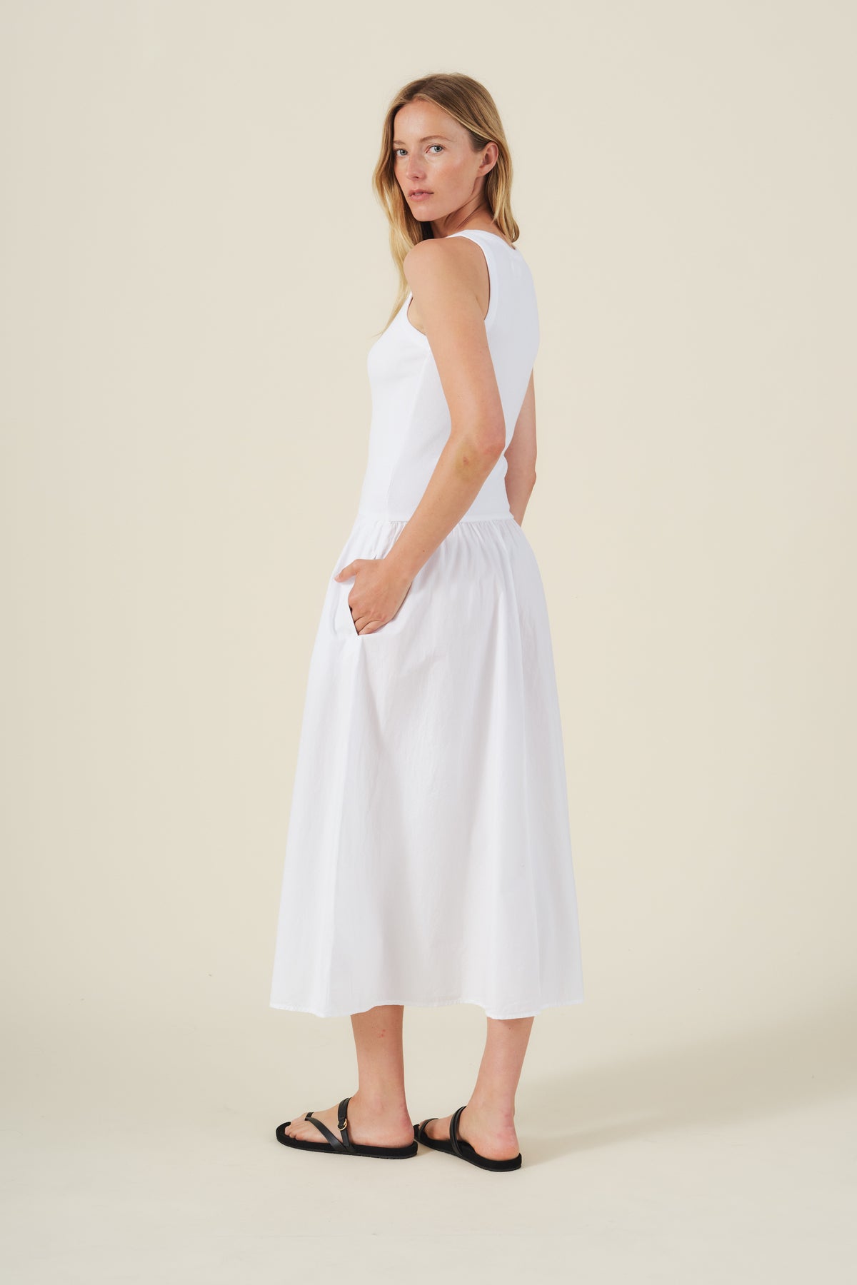 A woman stands sideways against a plain beige background wearing the ROMILLY DRESS by Velvet by Graham & Spencer—a sleeveless white ribbed bodice dress with pockets—paired with black sandals.
