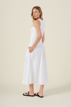 A woman stands sideways against a plain beige background wearing the ROMILLY DRESS by Velvet by Graham & Spencer—a sleeveless white ribbed bodice dress with pockets—paired with black sandals.