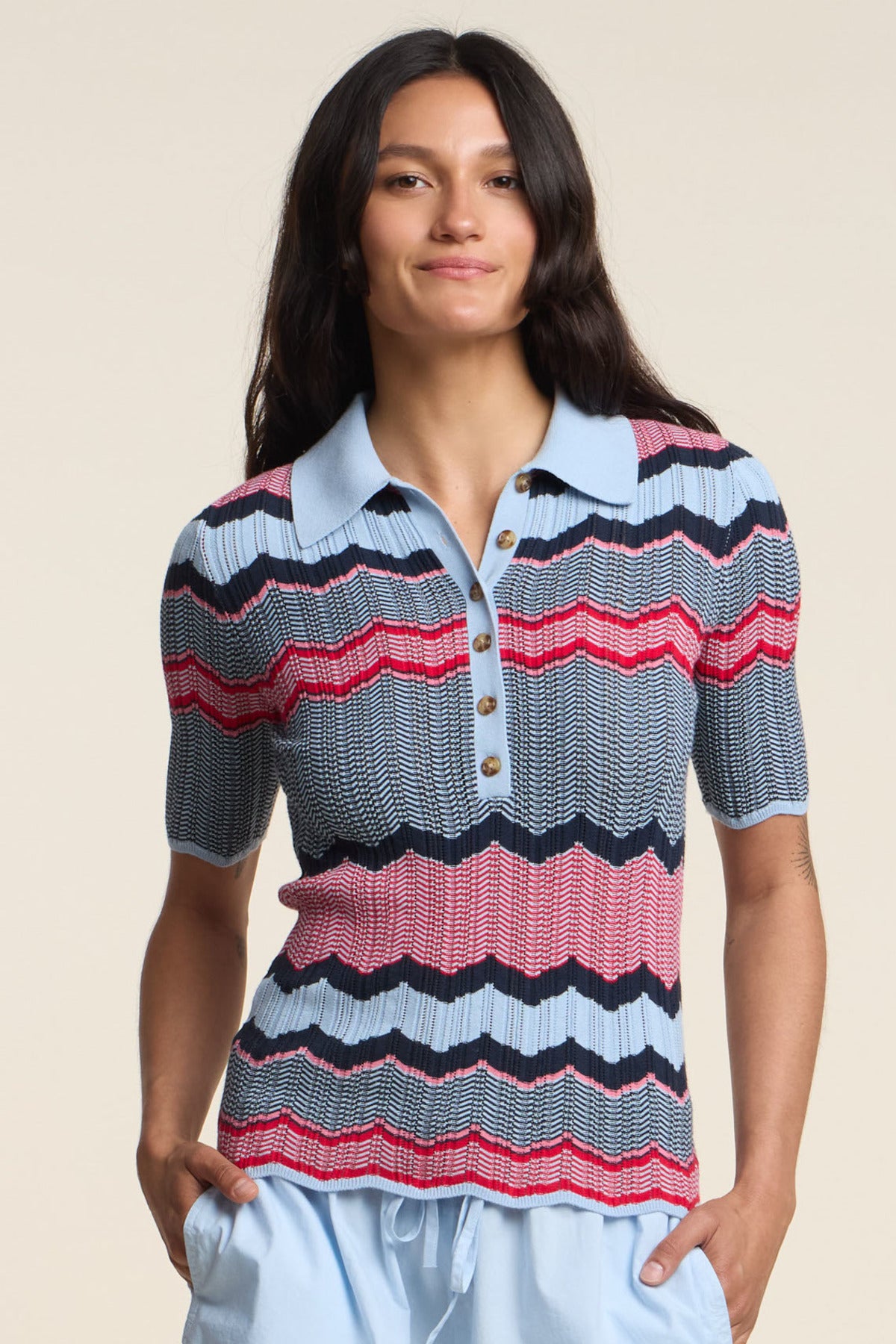 A woman faces forward against a beige background, wearing the fitted Velvet by Graham & Spencer ROSELLE POLO with red, navy, and light blue zigzag stripes, paired with light blue shorts.