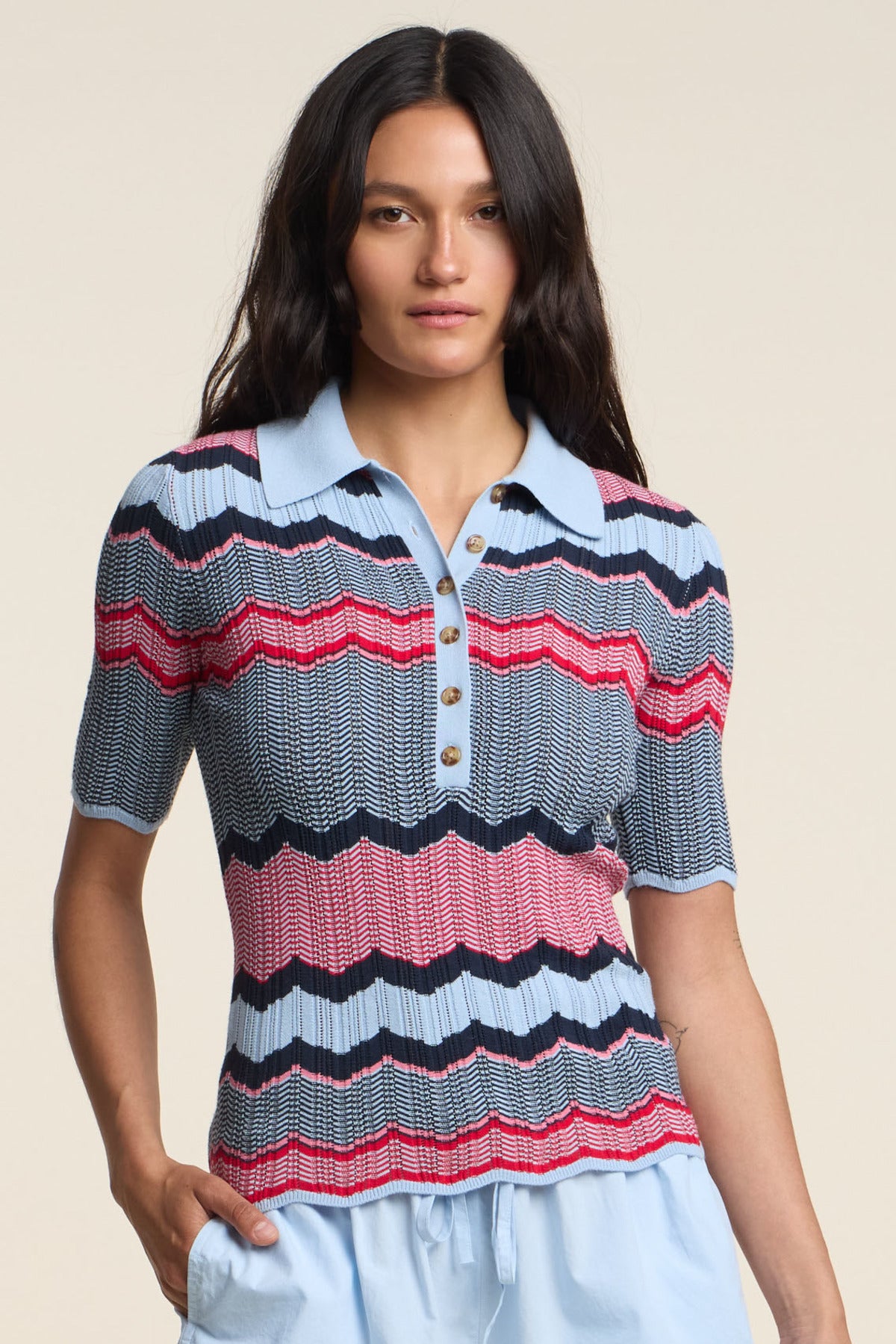 A woman wears the ROSELLE POLO by Velvet by Graham & Spencer, a short-sleeve button-up knit with blue, red, and black zigzag stripes, paired with light blue pants against a neutral background.