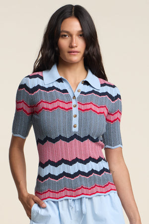 A woman wears the ROSELLE POLO by Velvet by Graham & Spencer, a short-sleeve button-up knit with blue, red, and black zigzag stripes, paired with light blue pants against a neutral background.