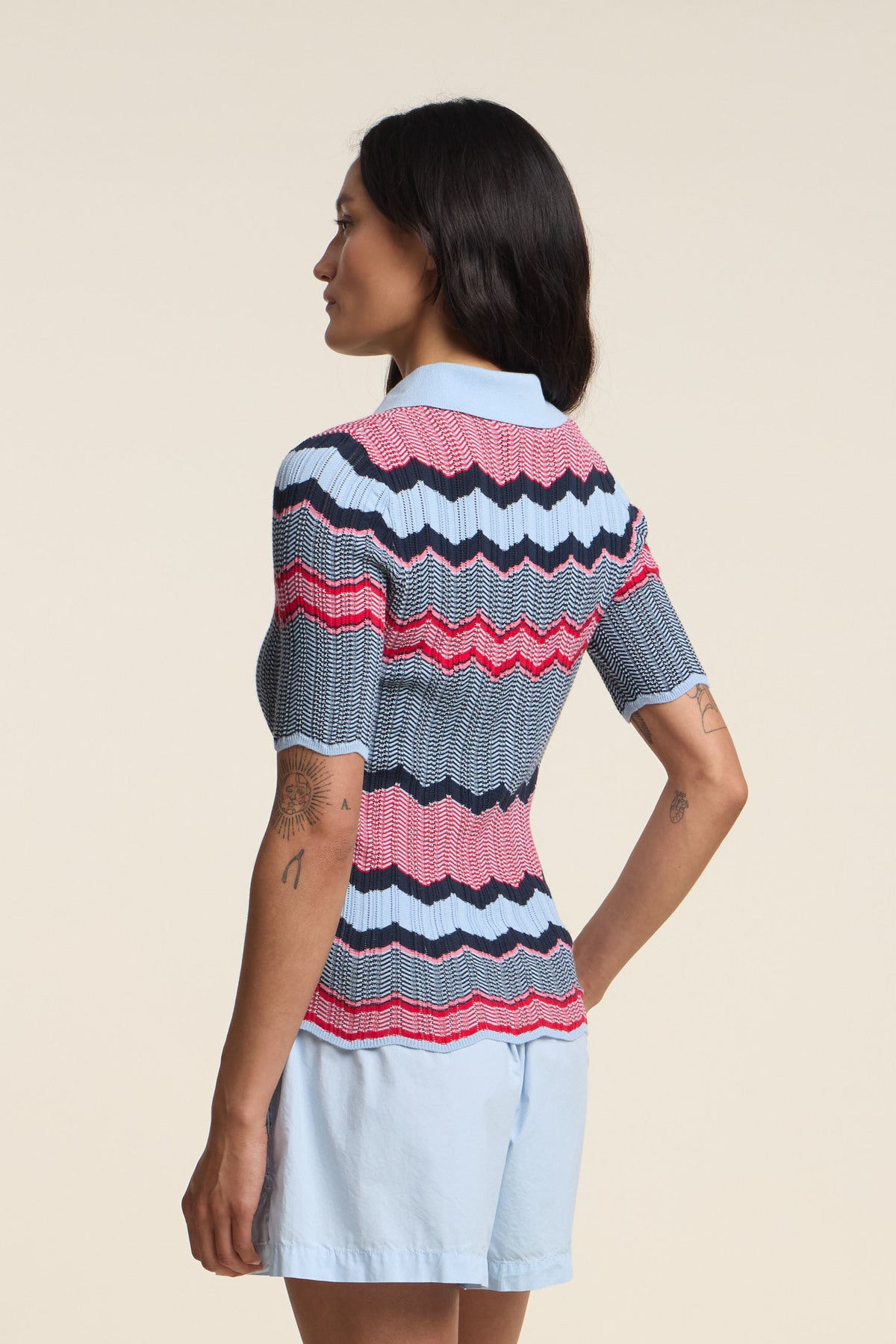 A woman faces away, wearing the Velvet by Graham & Spencer ROSELLE POLO with red, blue, and white zigzag stripes and a fitted silhouette, paired with light blue shorts.