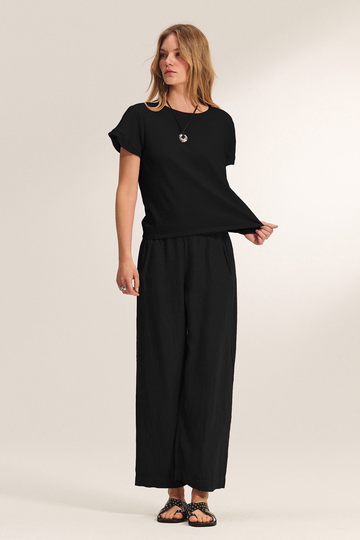 A woman wears the Velvet by Graham & Spencer LOLA LINEN PANT—a lightweight, elastic-waist, pull-on trouser—paired with a loose black t-shirt, sandals, and a necklace against a plain light background.-40659933200577