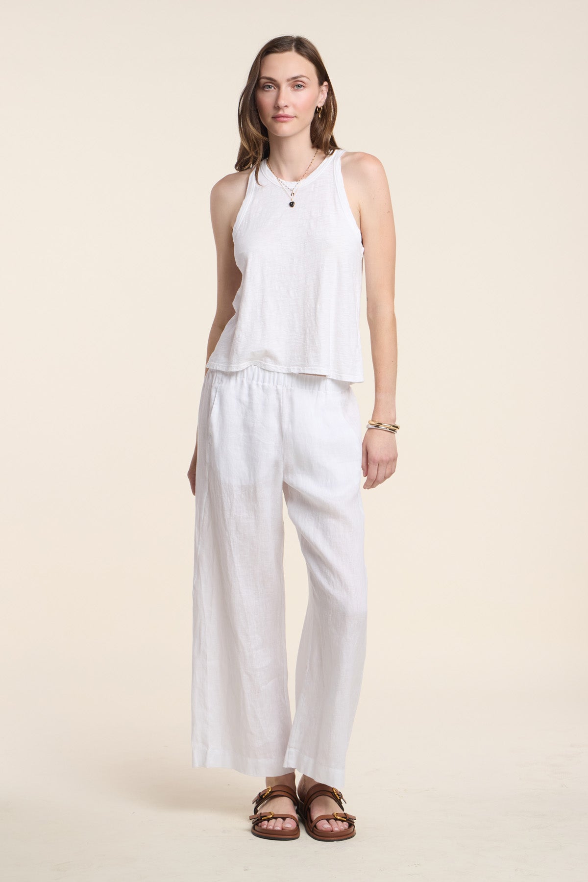 A woman faces forward in the SASHA TANK TOP by Velvet by Graham & Spencer, paired with white wide-leg pants, brown sandals, and simple jewelry, set against a plain beige background.