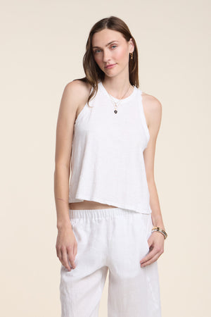 A woman stands against a plain background wearing the Velvet by Graham & Spencer SASHA TANK TOP—a relaxed fit white halter-style cotton slub tank—paired with white pants, bracelets, and layered necklaces.