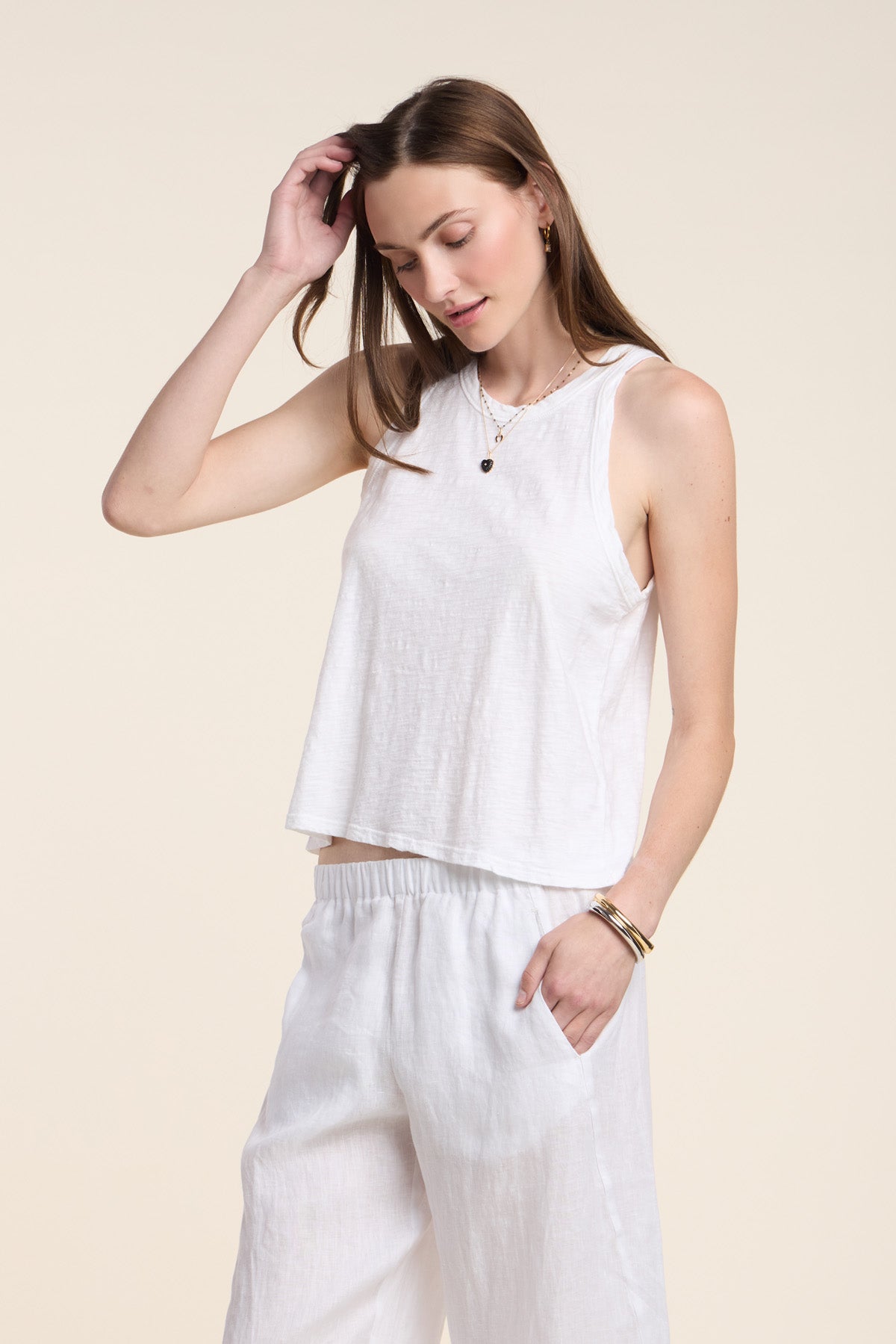 A woman wears the SASHA TANK TOP by Velvet by Graham & Spencer, styled with loose white pants, standing against a plain light background and touching her hair.