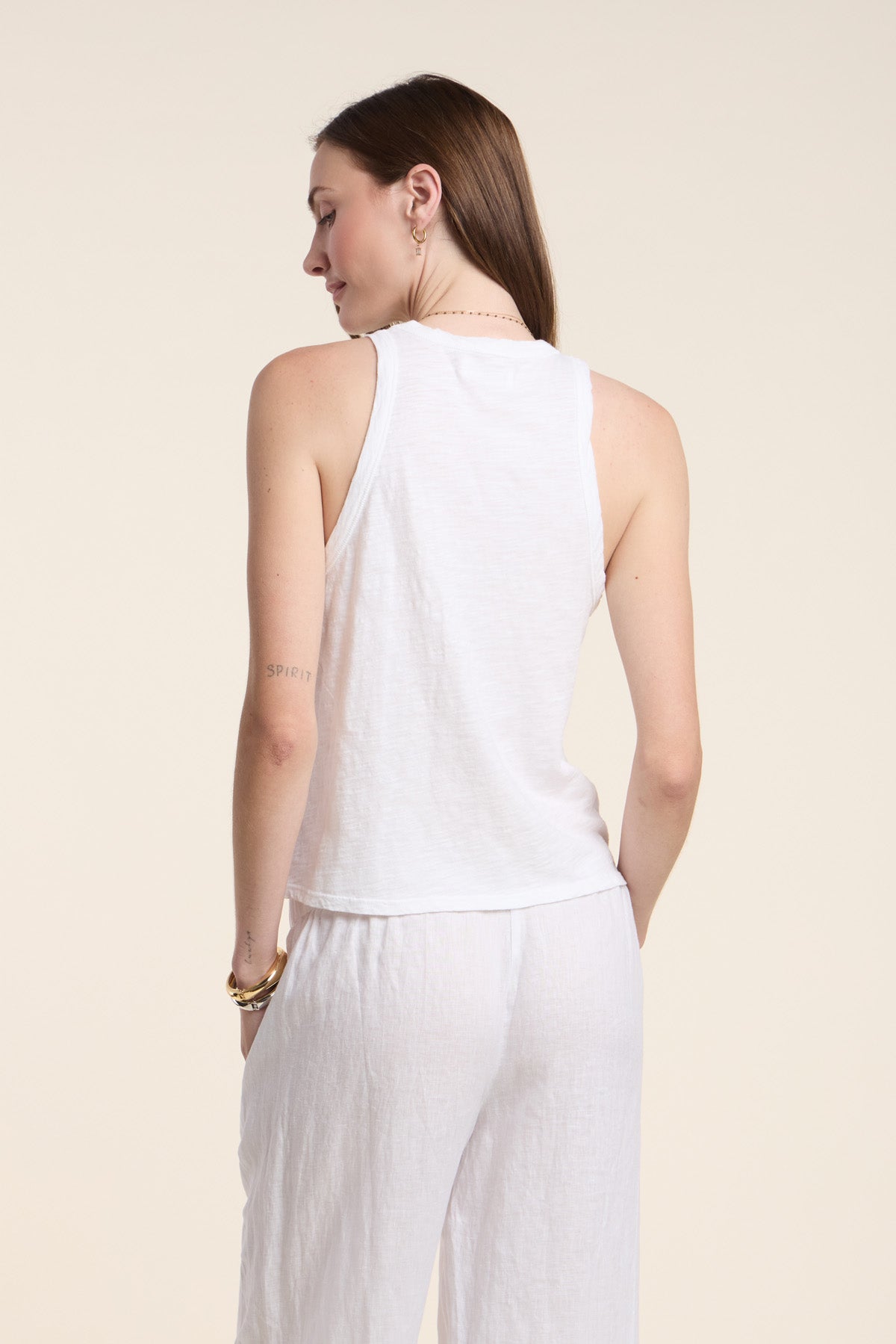A woman faces away, wearing the Velvet by Graham & Spencer SASHA TANK TOP in sleeveless white cotton slub with white pants. Her straight brown hair flows down her back, and a small tattoo is visible on her left arm.