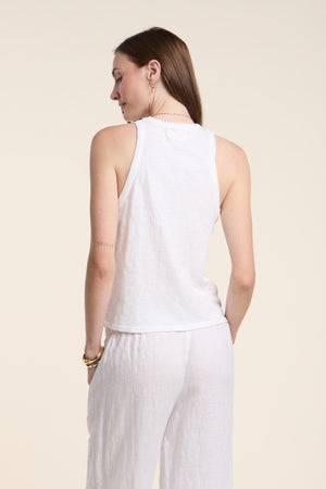 A woman faces away, wearing the Velvet by Graham & Spencer SASHA TANK TOP in sleeveless white cotton slub with white pants. Her straight brown hair flows down her back, and a small tattoo is visible on her left arm.
