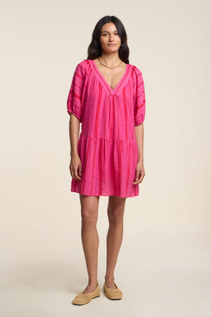 A woman stands against a plain background wearing the SELITA DRESS by Velvet by Graham & Spencer, a bright pink, short-sleeve mini dress with a V-neck, styled with beige woven flats.