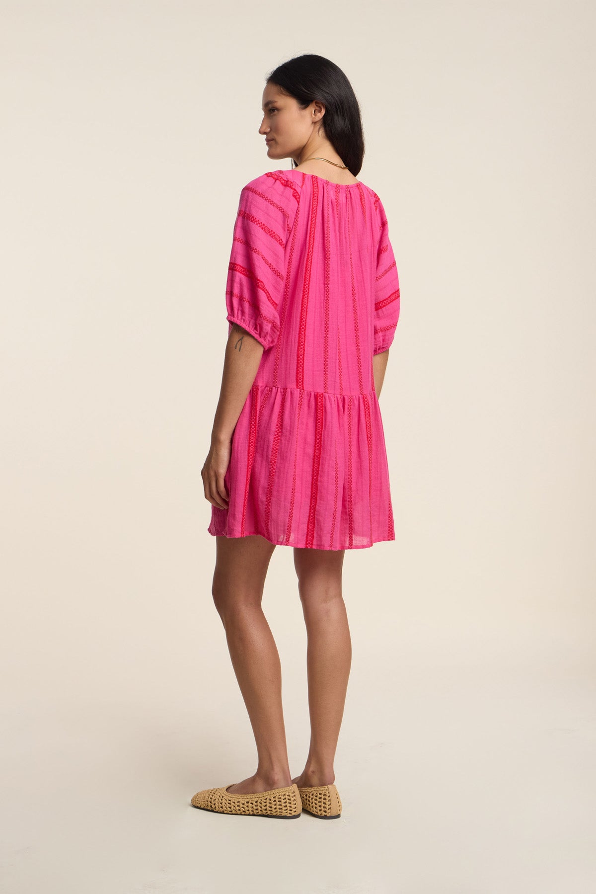 A woman faces sideways, wearing the SELITA DRESS by Velvet by Graham & Spencer—a loose geometric cotton jacquard pink mini dress with elbow-length puff sleeves—paired with beige woven slip-on shoes.-41372211445953