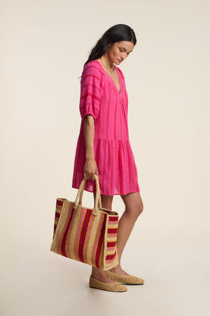 A woman wears the SELITA DRESS by Velvet by Graham & Spencer—a pink geometric cotton jacquard mini dress with statement puff sleeves—styled with a large striped woven tote and woven flats, standing against a plain light background.