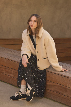 Wearing the ANIKA TEDDY JACKET by Velvet by Graham & Spencer over a black polka dot dress, a woman sits on a wooden bench and looks at the camera with a calm expression—effortlessly mastering cold-weather layering.
