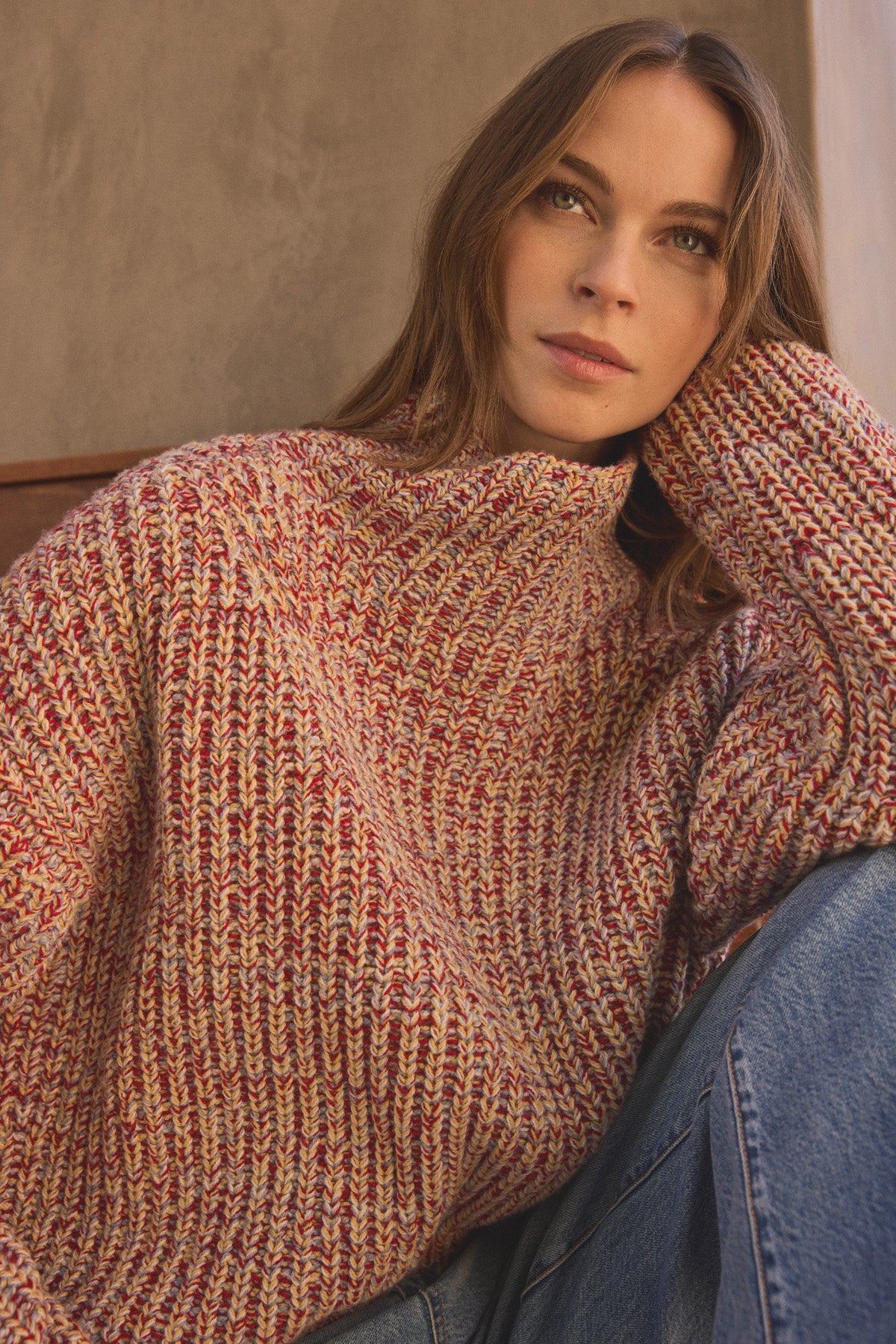 A woman with long brown hair wears the DENNIE SWEATER by Velvet by Graham & Spencer—a chunky wool blend sweater with a marled texture—paired with blue jeans. She sits with her head resting on her hand, looking slightly upward.